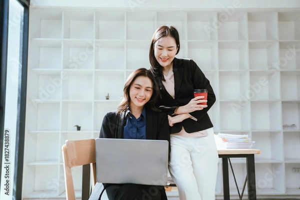 Obraz Two businesswomen work together at a laptop, smiling as they collaborate in an office with stacks of documents on the desk.