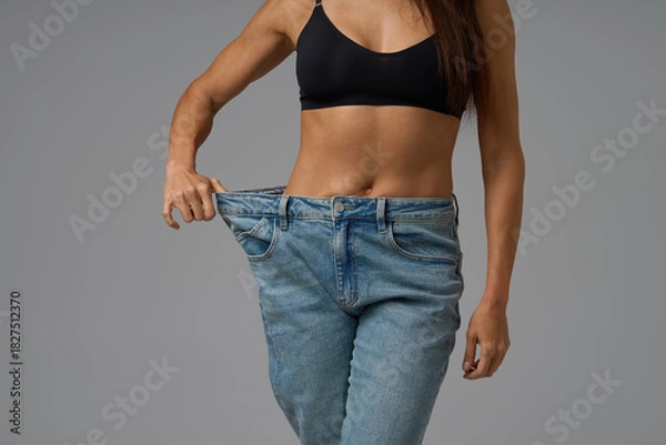 Obraz Fit woman in black sports bra pulling loose jeans to show weight loss progress on gray background