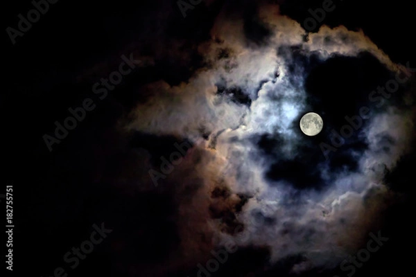 Fototapeta The moon illuminates the black sky around its light at night. Copy space for text. The moon behind the clouds.