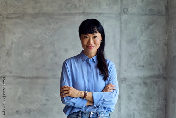 Fototapeta Asian Woman in Blue Shirt Stands Confidently With Crossed Arms in Modern Workspace