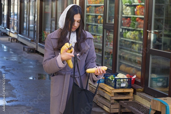 Obraz Young woman in a winter coat and headscarf looks into a shopping cotton bag while holding a bunch of bananas near an outdoor market with fruit crates, illustrating daily urban shopping.