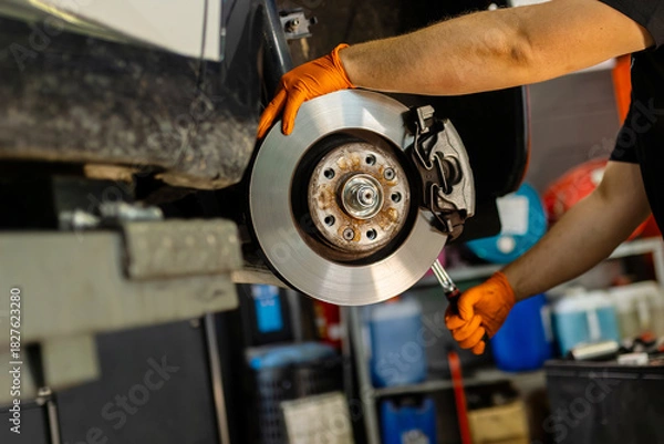 Fototapeta Mechanic hands with orange gloves installing or repairing a car brake disc. Maintenance of the braking system for vehicle safety in the garage.