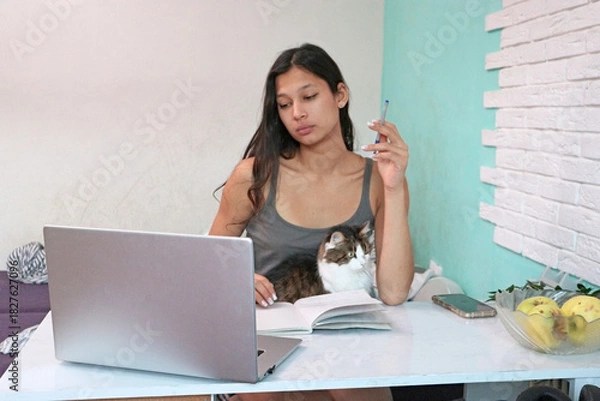 Fototapeta Young woman in a tank top sits at a white desk with a laptop, notebook, apples, and a cat on her lap, holding a pen and looking away. Suitable for study, pet, work from home, or cozy living stories.