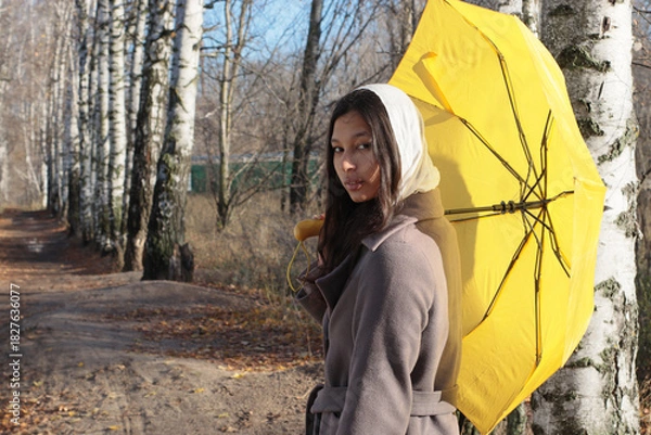 Obraz Young woman in a headscarf and brown coat stands on a forest path lined with birch trees, holding a yellow umbrella and looking at the camera. Suitable for autumn walk, beauty, seasonal content.