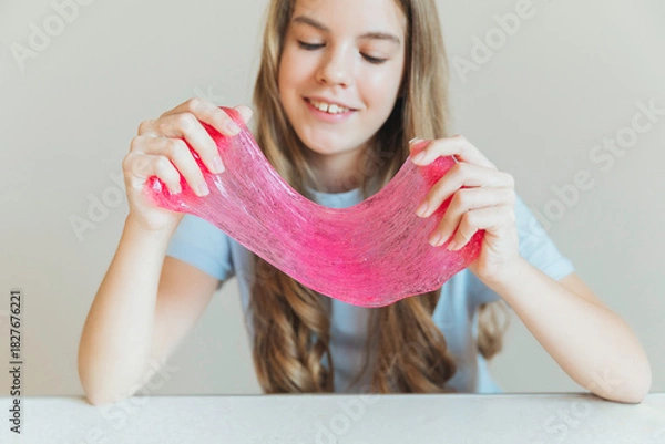 Fototapeta Close-up of hands stretching bright pink slime towards the camera, with a smiling girl in the background. Sensory toy