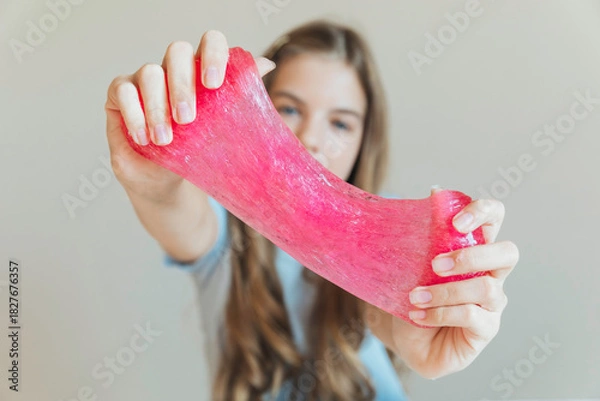 Fototapeta Close-up of hands stretching bright pink slime towards the camera, with a smiling girl in the background. Sensory toy