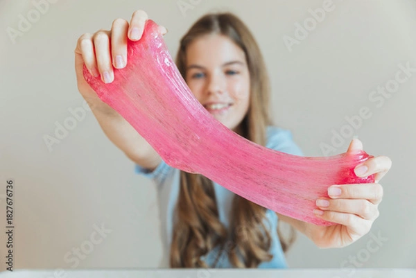 Fototapeta Close-up of hands stretching bright pink slime towards the camera, with a smiling girl in the background. Sensory toy