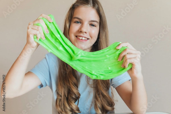 Fototapeta Smiling girl stretches wide a blob of vibrant neon green slime. Fun sensory toy and creative anti-stress activity