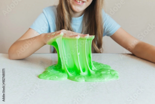 Fototapeta Smiling girl holding and stretching lime green slime wide, showcasing its elasticity and stringy texture. Fun sensory play