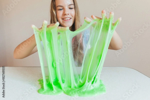 Fototapeta Smiling girl holding and stretching lime green slime wide, showcasing its elasticity and stringy texture. Fun sensory play