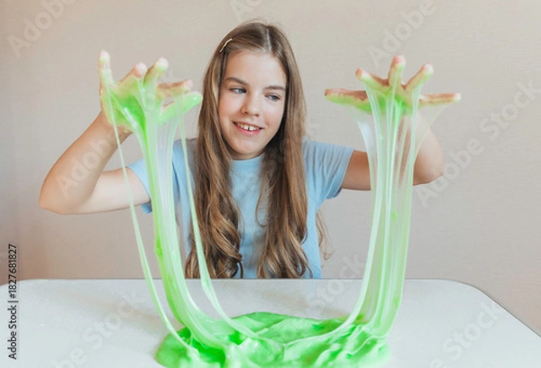 Fototapeta Smiling girl holding and stretching lime green slime wide, showcasing its elasticity and stringy texture. Fun sensory play