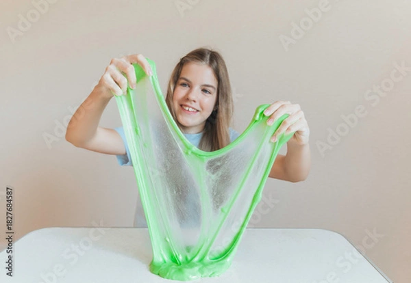 Fototapeta Girl stretching bright neon green slime into a thin translucent sheet on the table. Fun sensory and creative DIY hobby
