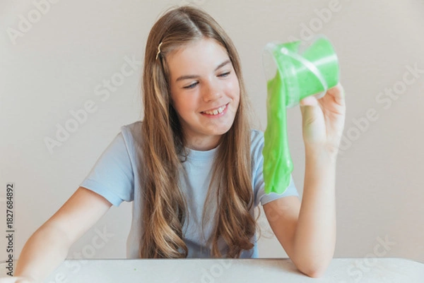Fototapeta Smiling girl pouring vibrant green slime from a container onto the table. Trendy DIY and fun sensory play activity