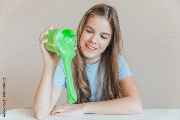 Fototapeta Smiling girl pouring vibrant green slime from a container onto the table. Trendy DIY and fun sensory play activity