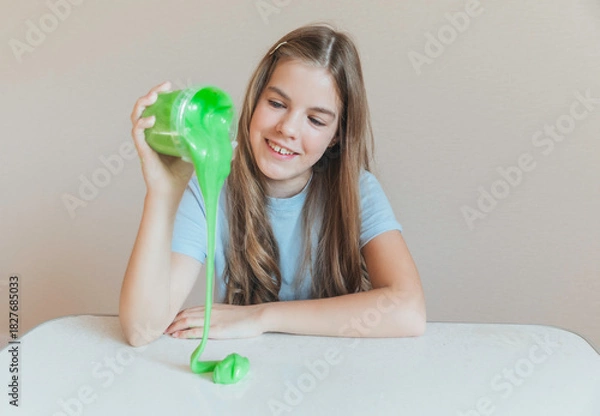 Fototapeta Smiling girl pouring vibrant green slime from a container onto the table. Trendy DIY and fun sensory play activity