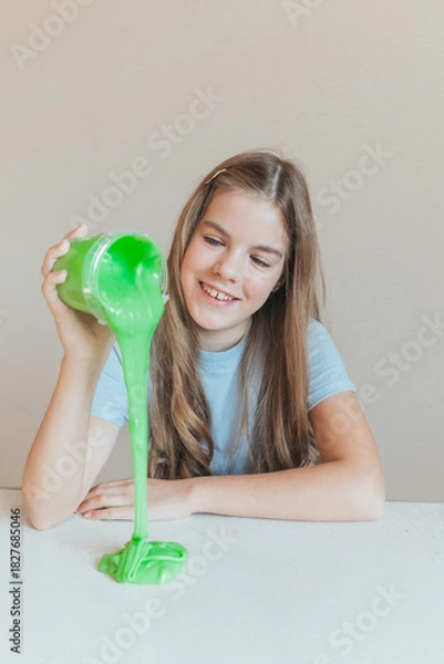 Fototapeta Smiling girl pouring vibrant green slime from a container onto the table. Trendy DIY and fun sensory play activity