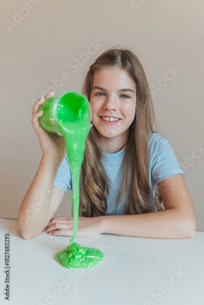 Fototapeta Smiling girl pouring vibrant green slime from a container onto the table. Trendy DIY and fun sensory play activity