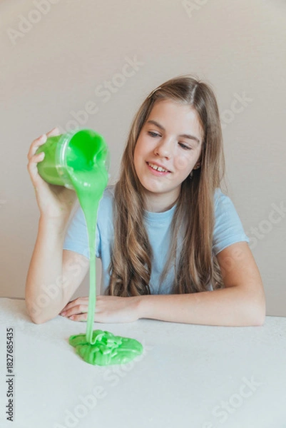 Fototapeta Smiling girl pouring vibrant green slime from a container onto the table. Trendy DIY and fun sensory play activity