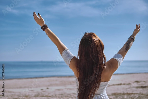 Fototapeta Rear view of young woman in white dress stands on sunny beach with her arms raised toward sky, feeling freedom and tranquillity of ocean. Deep connection with nature and inner balance concept