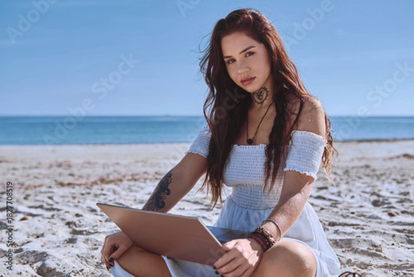 Fototapeta Young 20s beautiful freelancer woman in white summer dress sitting on sandy beach with laptop, posing, looking at camera. Teleworking, online studying, blogging concept