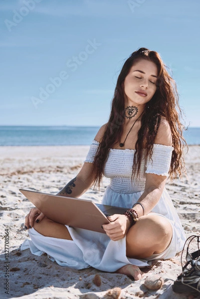 Fototapeta Young woman in white summer dress sits on the beach with laptop with eyes closed, relaxing, enjoy summer sun, distracted from freelance work or studying process on nature. Freelance, modern tech use