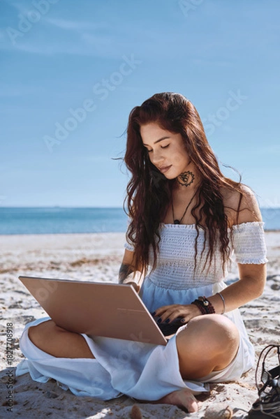 Fototapeta Young woman in summer dress sits on sandy beach with laptop, looks at screen and typing, chatting, engage in freelance work or studying process on nature. Telework, modern tech use, blogging concept