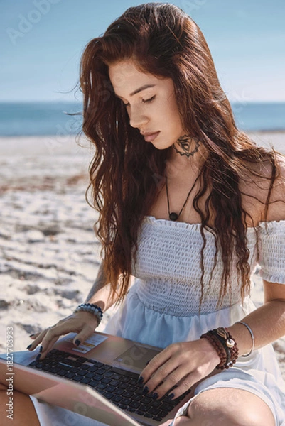 Fototapeta Young woman in summer dress sits on sandy beach with laptop, looks at screen and typing, chatting, engage in freelance work or studying process on nature. Telework, modern tech use, blogging concept