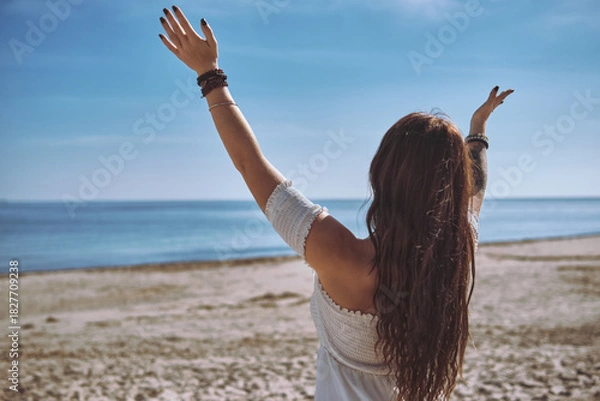 Fototapeta Rear view of young woman raises her hands in serene gesture of mindfulness while facing calm blue ocean on the beach. Freedom, spiritual renewal by ocean
