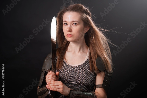 Fototapeta Portrait of a young woman, fantasy warrior in armor with a sword in her hands isolated on a dark background.