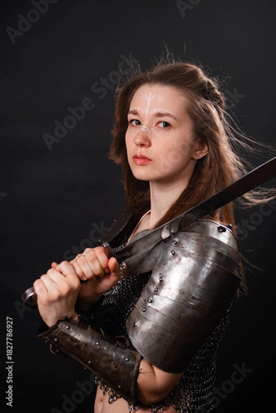 Fototapeta Portrait of a young woman, fantasy warrior in armor with a sword in her hands isolated on a dark background.