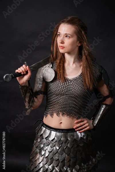Fototapeta Portrait of a young woman, fantasy warrior in armor with a sword in her hands isolated on a dark background.