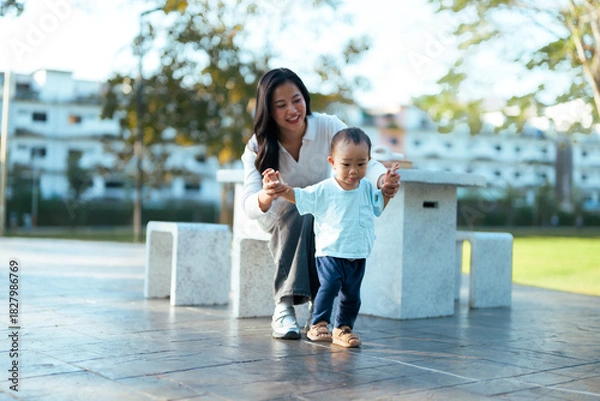 Fototapeta Mother supporting toddler learning first steps in park