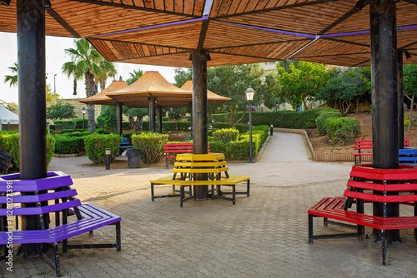 Obraz Bright circular benches painted in rainbow colors beneath umbrella-shaped wooden canopies in a public park. Artistic urban seating surrounded by greenery and walking paths.