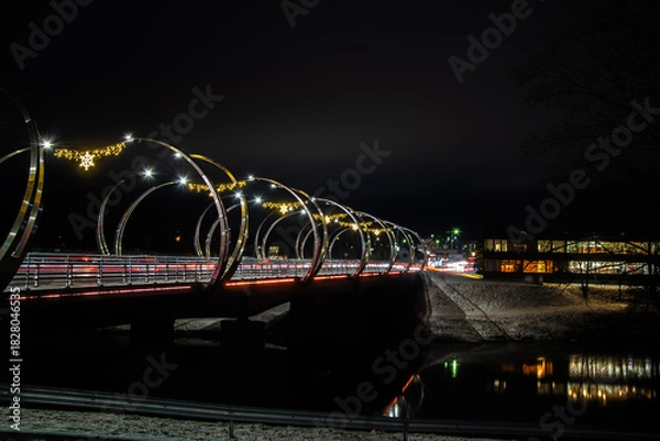 Obraz Night long exposure of a modern bridge with festive lights and traffic streaks over a dark river, winter banks and city glow.
