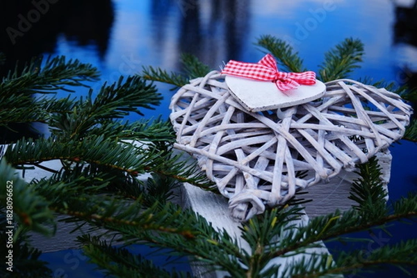 Obraz Christmas decorations made of natural materials. On the big heart is a small heart with a red and white ribbon. Against the background - Christmas tree branches and blue water surface.