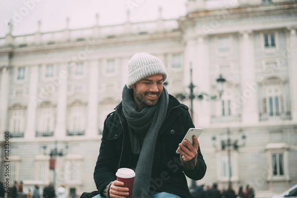 Fototapeta Young blonde man on the mobile phone and drinking coffee near the royal palace in winter  .
