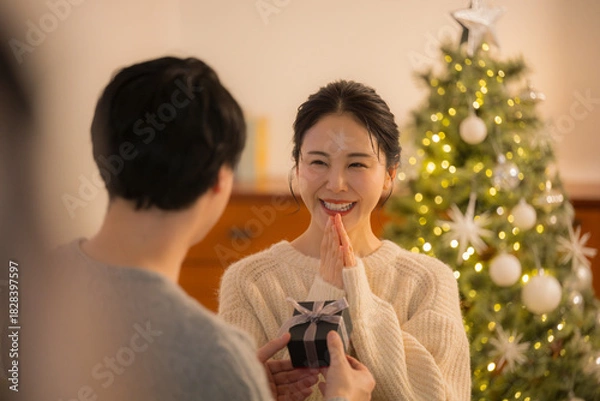 Obraz クリスマスツリー前でプレゼントを受け取り喜ぶ女性　カップルの贈り物シーンの冬イベントイメージ