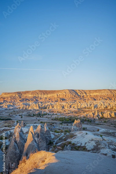 Fototapeta A striking vertical photograph of Cappadocia’s iconic rock formations illuminated by warm sunrise light in Turkey. The golden cliffs, dramatic fairy chimneys, and expansive blue sky