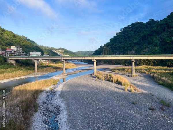 Fototapeta Early morning view of elevated bridge spanning wide rocky riverbed with shallow stream in Wulai, New Taipei City, Taiwan. Golden light on lush green mountains.