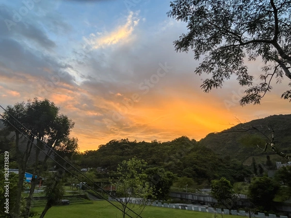 Fototapeta Vivid orange and yellow sunrise sky above the lush green mountains and river area in Wulai, New Taipei City, Taiwan, seen from a park or open space.