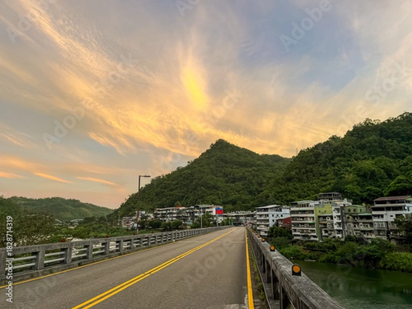 Fototapeta Empty bridge road leading into the village houses in Wulai, New Taipei City, Taiwan, set against a stunning golden and orange sunrise sky over lush green mountains.