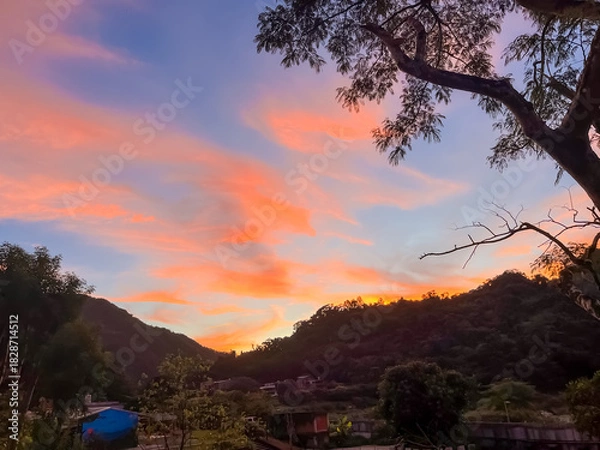 Obraz Breathtaking pink and orange clouds against a blue sky over the lush green hills and town of Wulai, New Taipei City, Taiwan, framed by a tree branch silhouette.