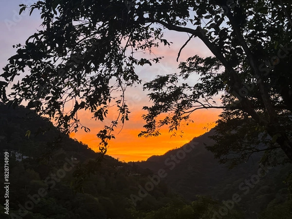 Fototapeta Striking orange and yellow sunrise illuminates the sky above a dark mountain valley silhouette in Wulai, New Taipei City, Taiwan, creating a dramatic and powerful scene.