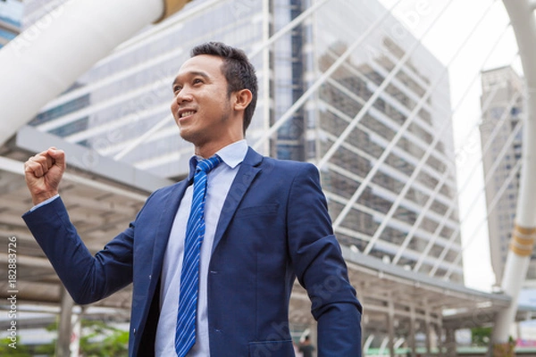 Obraz Businessman smiling and raising his fist in the air, with office building background - business success, achievement, and win concepts
