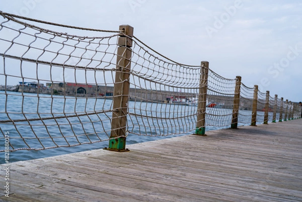 Fototapeta Wooden Boardwalk with Rope Net Fence by the Water on a Cloudy Day