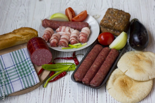 Fototapeta sausages, bacon, vegetables and bread on white wooden table