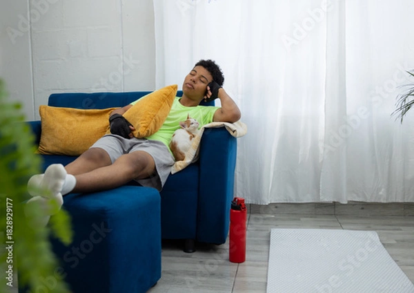 Obraz Brown skinned teenager asleep on the couch after his home workout, while his cat rests beside him. A warm scene that reflects relaxation after exercising at home