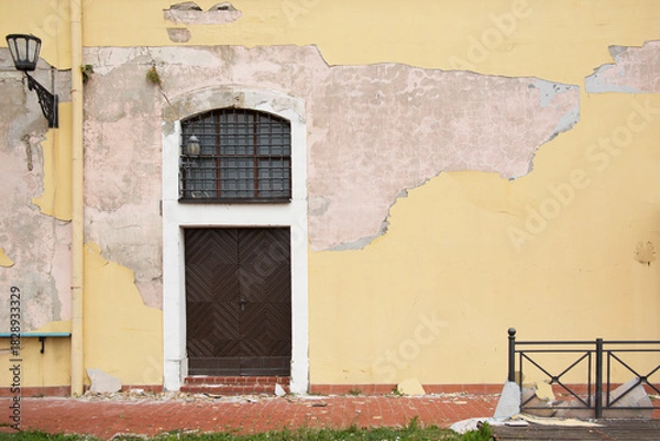 Fototapeta A yellow wall of an old building with a brown door and crumbling plaster