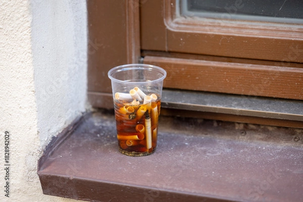 Obraz A plastic cup filled with cigarette butts soaked in liquid, left on a window ledge outdoors, showing urban littering and pollution.