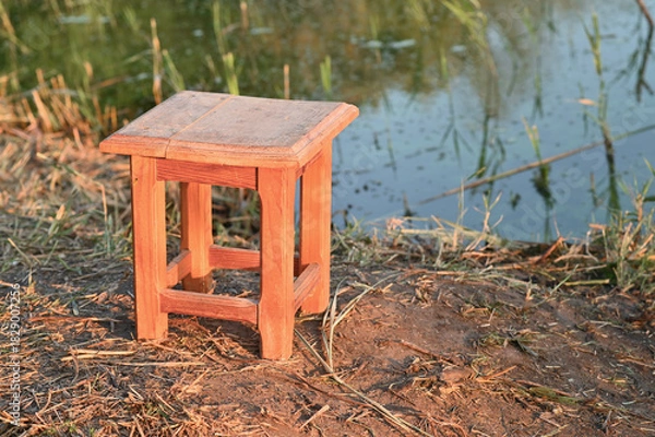 Fototapeta Small wooden stool stands on dry ground near calm water edge under warm sunlight. rustic handmade furniture placed in quiet natural environment. peaceful countryside atmosphere.